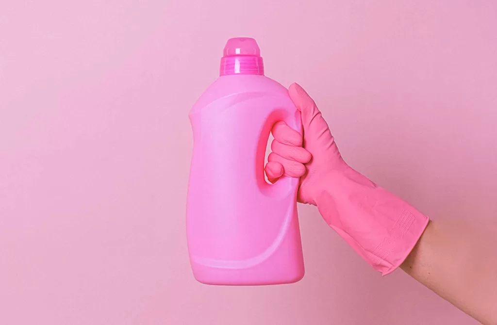 Hand with a pink glove on it holding a pink bottle of laundry detergent against a pink wall.