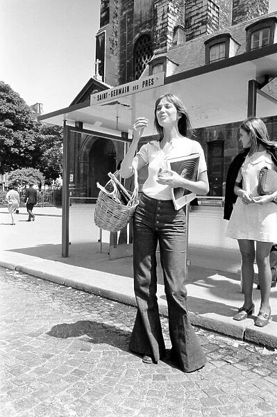 Streetstyle shot of actress Jane Birkin wearing denim flare leg jeans and carrying a basket, posing on the streets in France.