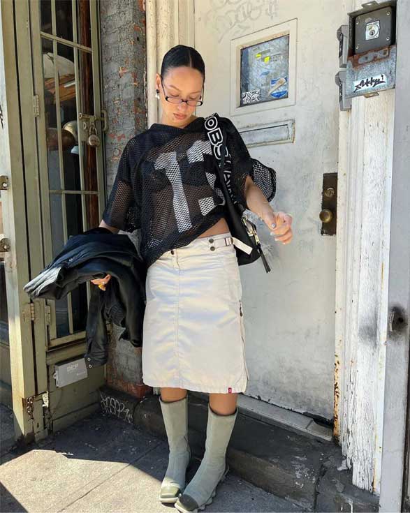 Young Gen-Z woman in an eclectic thrifted outfit: sheer mesh jersey shirt, long cargo skirt, tall boots and small miu miu glasses