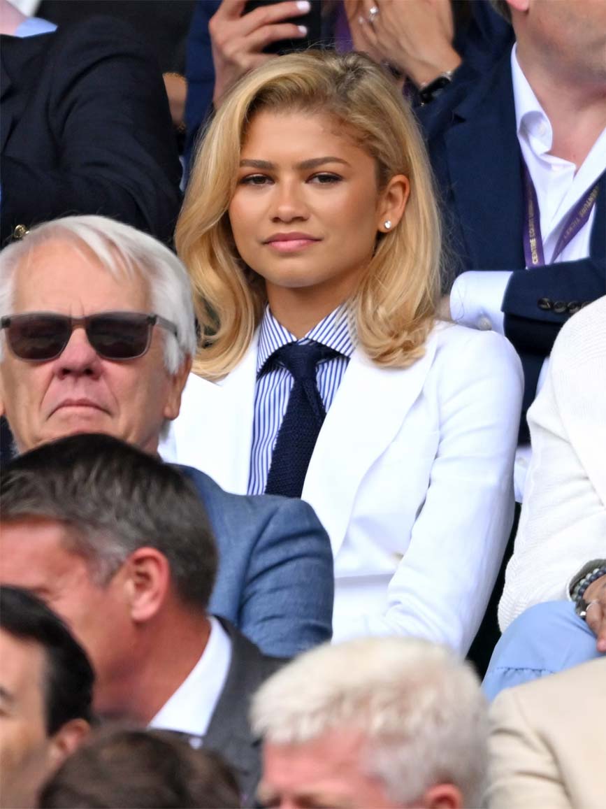 Zendaya at Wimbledon wearing a white blazer, white and blue button down pinstripe shirt, and a navy necktie with blonde hair